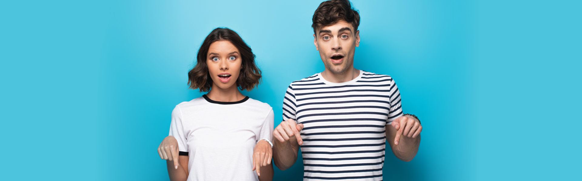 surprised man and woman pointing down with fingers and looking at camera on blue background